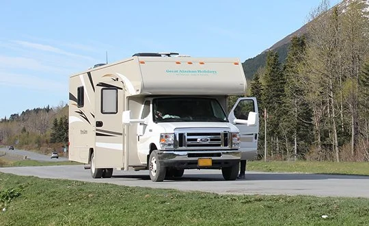 White Class C motorhome RV driving on mountain highway with forest and hills in background during daytime.